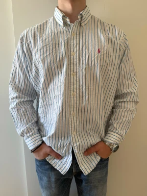 Randig skjorta från Ralph Lauren - Klassisk vit skjorta med blå ränder från Ralph Lauren. Skjortan har lång ärm, knappar framtill och en liten rosa logga broderad på bröstet. Tillverkad i bomull och har en normal passform, storlek XL i mycket bra skick utan defekter
