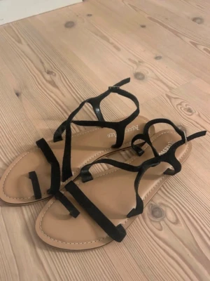 Svarta remsandaler från Accessorize - Säljer ett par stilrena sandaler från Accessorize med tunna svarta remmar i mockaimitation och beige sula. Sandalerna har öppen tå och justerbara remmar runt ankeln för en snygg och bekväm passform. Perfekta till sommaren och lätta att matcha.