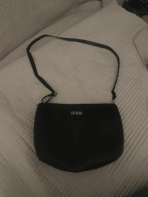 Svart axelväska från Guess - Snygg svart axelväska från Guess med justerbar axelrem och stilren design. Väskan har en slät, lätt strukturerad yta i konstläder och ett diskret Guess-logo i silver framtill. Perfekt för dig som vill ha en enkel men trendig väska till vardagslooken.