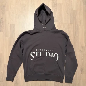 Mörkgrå hoodie från Jack & Jones - Säljer en snygg mörkgrå hoodie från Jack & Jones Originals med stor vit STUDIO-print på framsidan. Tröjan har huva med tryckknappar, magficka och mjukt fleecefoder på insidan. 