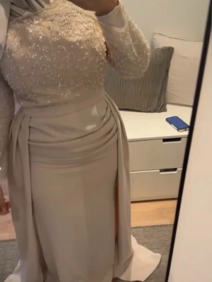Festklänning från Jolie bridal - Elegant långklänning i begie färg från Jolie bridal. Klänningen har glittriga, pärlbroderade långärmar och en mjukt draperad kjol. Överdelen är dekorerad med pärlor och paljetter, vilket ger en lyxig känsla. Perfekt för bal eller festliga tillfällen. Det står att den är xxl men den passar en ma-l. Använd bara en gång 