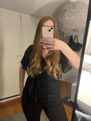 Svart omlottblus med knytband - Säljer en svart omlottblus med knytband i midjan och v-ringning. Blusen har korta, transparenta ärmar och är tillverkad i ett lätt, syntetiskt material. Perfekt för dig som gillar en stilren och trendig look.