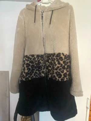 Teddyjacka med leopardmönster och huva - Mysig teddyjacka i beige, svart och leopardmönstrad panel. Jackan har dragkedja framtill, huva och långa ärmar. Perfekt för dig som vill sticka ut med en cool och trendig look. Tillverkad i fluffigt teddy-material för extra komfort.