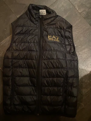 Svart dunväst från Emporio Armani - Snygg svart dunväst från Emporio Armani EA7 med guldfärgad logga på bröstet. Västen har hög krage, dragkedja framtill och quiltad design. Perfekt för lager-på-lager och ger en sportig vibe till din outfit.