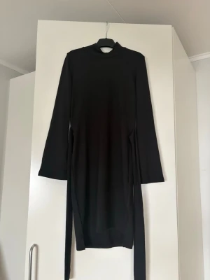 Svart långärmad klänning med knyt - En svart, långärmad klänning med hög hals och knytband i midjan som ger en snygg siluett. Klänningen har en enkel och stilren design, perfekt för dig som gillar minimalistiskt mode. Materialet känns mjukt och stretchigt, troligen bomull eller polyester.