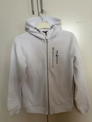 Vit hoodie från Sail Racing - Säljer en vit hoodie från Sail Racing med dragkedja framtill och en extra bröstficka med zip. Hoodien har en broderad logga på bröstet och är tillverkad i mjukt material. Perfekt för en clean och sportig look.