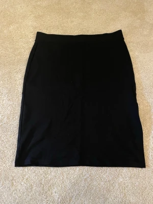 Svart enkel kjol från KappAhl - Klassisk svart kjol från KappAhl i rak modell. Kjolen är knälång och har en stilren design utan detaljer eller mönster, vilket gör den lätt att matcha med olika toppar. Perfekt basplagg i garderoben.