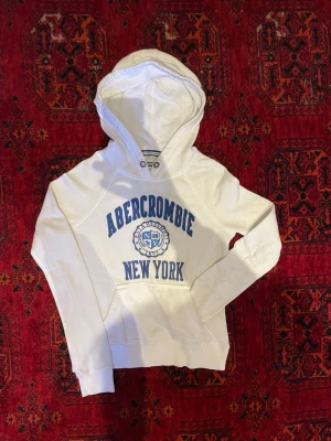 Abercrombie & Fitch Hoodie - Snygg vit hoodie från Abercrombie med blå text och emblem på bröstet där det står 'Abercrombie New York'. Klassisk känguruficka framtill och huva med dragsko. Mjuk bomullsblandning som känns skön mot huden. Undrar du något är det bara att skriva😁🙌