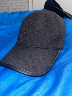 Svart keps - Säljer en svart keps med det klassiska monogrammönstret över hela kepsen. Kepsen har böjd skärm med svart kant i läderimitation och är tillverkad i tyg. Perfekt för dig som vill ha en stilren och exklusiv accessoar.