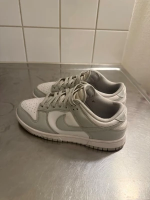 Nike Dunk Low vit/grå sneakers - Nike Dunk Low sneakers i vitt och ljusgrått läder med klassisk swoosh på sidan. Lågt skaft, perforerad tåbox och platt sula. Broderad Nike-logga på hälen och matchande vita skosnören. Perfekt för dig som gillar stilrena och tidlösa sneakers.