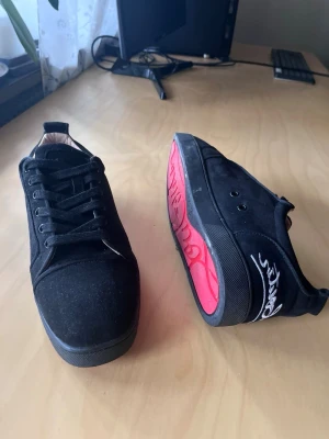 Christian Louboutin skor  - Snygga svarta sneakers med låg profil och svart mockaliknande material. Skorna har svarta skosnören och en unik röd sula med vit text. Perfekta för dig som vill ha en clean och stilren look med en twist under foten. Storlek 44. Bra skick!