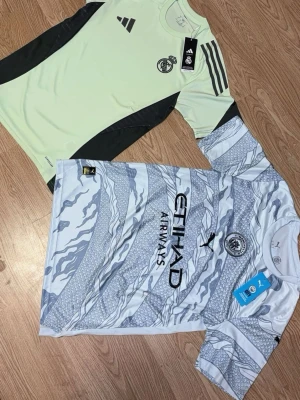 Manchester City & Real Madrid matchtröjor Pumpa & Adidas - Säljer Manchester City och Real Madrid matchtröjor, en från Puma i vitt och ljusgrått med grafiskt mönster och Etihad Airways-tryck, samt en Real Madrid tröja i ljusgrönt med svarta detaljer och klubbmärke på bröstet. Perfekta för fotbollsträning eller att bära till vardags.