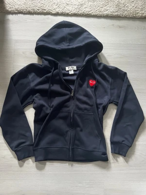 Mörkblå hoodie Comme des Garçons Play - Mörkblå hoodie med dragkedja från Comme des Garçons Play. Klassisk modell med huva och fickor framtill. På bröstet sitter det ikoniska röda hjärtat med ögon. Tillverkad i mjukt bomullsmaterial och har ribbade muddar vid ärmslut och nederkant.