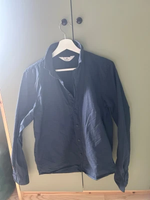 Mörkblå skjorta från H&M - Snygg mörkblå skjorta från H&M med klassisk krage och knäppning framtill. Skjortan har långa ärmar och är tillverkad i mjuk bomull som känns skön mot huden. Perfekt för en stilren och enkel look.