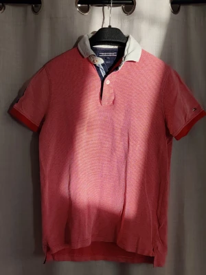 Rosa piké från Tommy Hilfiger (slimfit) - Säljer en rosa piké från Tommy Hilfiger med vit krage och knappslå. Tröjan har korta ärmar och diskret logga på ärmen. Slim fit-modell i bomull som ger en fräsch och sportig look. Perfekt för dig som gillar klassisk stil med en twist.