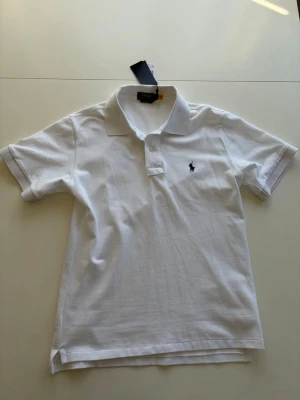Vit pikétröja från Polo Ralph Lauren - Klassisk vit pikétröja från Polo Ralph Lauren. Tröjan har krage, korta ärmar och knappar vid halsen. Modellen är custom slim fit och tillverkad i bomull för en skön känsla. ALDRIG ANVÄND. Säljer för jag fick den i present och har en likadan! Pris 699kr