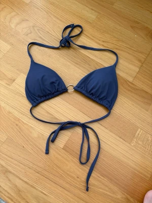 Mörkblå bikiniöverdel , subdued, med ringdetalj - Snygg mörkblå bikiniöverdel med trekantskupor och en silverfärgad ringdetalj i mitten. Knytband i nacken och runt ryggen för justerbar passform. Perfekt för strandhäng eller pooldagar.