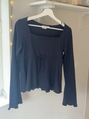 Mörkblå långärmad topp Gina Tricot - Mörkblå långärmad topp från Gina Tricot med fyrkantig ringning och liten knytning i midjan. Toppen har lätt utsvängda ärmslut och vågig kant nertill, vilket ger en söt och trendig look. Perfekt att styla med jeans eller kjol.