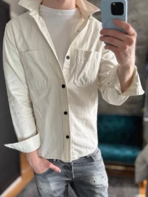 Vit overshirt med svarta knappar - Stilren vit overshirt med svarta knappar och två bröstfickor. Overshirten har en avslappnad passform och är tillverkad i ett kraftigare bomullstyg. Perfekt att bära öppen över en t-shirt för en casual look.