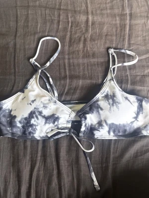 Svartvit bikiniöverdel med knyt - Säljer en snygg bikiniöverdel i svartvitt batikmönster. Modellen har smala axelband med justerbar längd och knyt framtill för extra detalj. Materialet känns mjukt och stretchigt, perfekt för dagar på stranden.