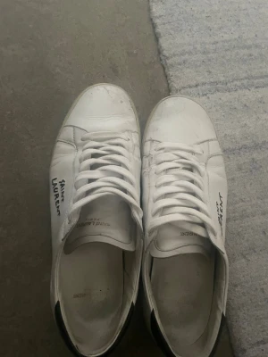 Vita Saint Laurent sneakers - Säljer ett par klassiska vita sneakers från Saint Laurent med handskriven logga på sidan. Skorna har låga skaft, vita snören och beige sula. Tillverkade i läder med stilren och minimalistisk design som passar till det mesta.