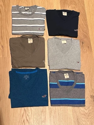 Mix av Hollister t-shirts, V-ringad - Säljer ett paket med sex snygga t-shirts för herr från Hollister. Alla har V-ringning och korta ärmar. Färgerna varierar mellan marinblå, grå, brun, blå och randiga varianter i grått och blått. Materialet är mjuk bomull och varje t-shirt har Hollisters klassiska logga broderad på bröstet.