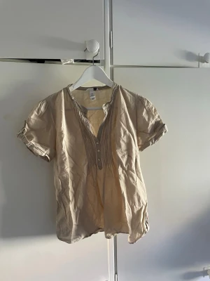 Beige blus från H&M med knappar - Luftig beige blus från H&M med korta ärmar och dekorativa knappar framtill. Blusen har fina veckdetaljer längs knappraden och en avslappnad passform. Tillverkad i ett tunt, mjukt material som känns skönt mot huden.