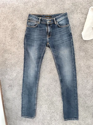 Blå skinny jeans från Nudie Jeans - Säljer ett par blå skinny jeans från Nudie Jeans med klassisk femficksmodell och snygga slitningar. Jeansen har normal midja, smal passform och är tillverkade i stretchigt denim för extra komfort. Perfekta för en avslappnad och trendig look.