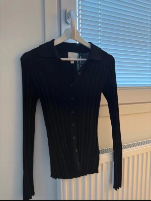 Svart ribbad kofta med knappar - Säljer en svart ribbad kofta med långa ärmar och knäppning framtill. Koftan har en klassisk krage och är figurnära i modellen, perfekt att styla till olika outfits. Materialet är stretchigt och mjukt för en bekväm känsla.