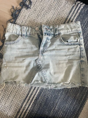 Ljus jeanskjol med rå kant - Säljer en ljusblå jeanskjol med rå, fransig kant och slitna detaljer. Kjolen har klassiska fem fickor, bälteshällor och snygga broderade bakfickor. Perfekt för dig som gillar en avslappnad och trendig stil.