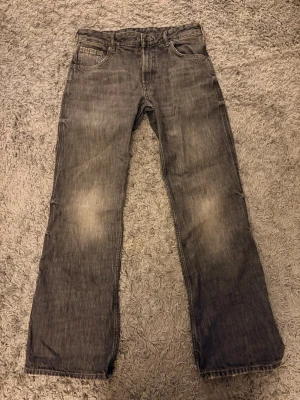 Grå raka jeans med slitningar - Säljer ett par grå jeans med raka ben och diskreta slitningar på låren. Klassisk femficksmodell med knapp och dragkedja framtill. Jeansen har en tvättad look och är tillverkade i denim, perfekt för en avslappnad stil.