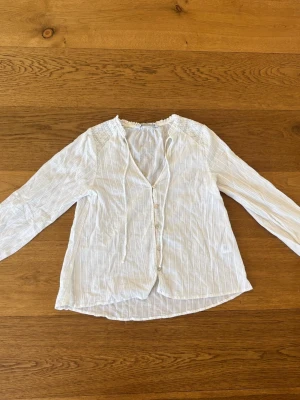 Vit boho-blus med knappar Zara - Luftig vit blus i bohostil med tunna vertikala ränder och dekorativa knappar framtill. Blusen har långa ärmar med knapp vid ärmslut och snörning vid halsen. Materialet är lätt och krinklat, perfekt för en avslappnad look.