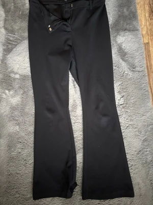Svarta bootcut kostymbyxor L/30 - Snygga svarta kostymbyxor med bootcut-modell och hög midja. Byxorna har hällor för skärp och stängs med hake och dragkedja. Perfekta för en clean och stilren look. Materialet är mjukt och följsamt för extra komfort. 