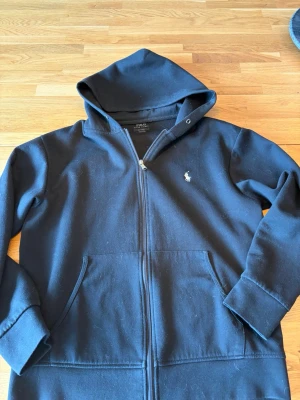 Svart zip up från Polo Ralph Lauren - Svart hoodie med dragkedja från Polo Ralph Lauren. Klassisk design med huva, fickor framtill och den ikoniska vita logon broderad på bröstet. Tillverkad i mjukt bomullsmaterial som är skönt att ha på sig. Perfekt för en avslappnad och stilren look.