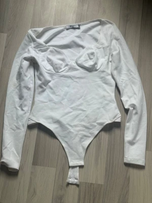 Vit långärmad body från NAKD - Trendig vit body från NAKD med långa ärmar och markerade kupor. Tillverkad i mjukt material för en bekväm passform. Perfekt att styla med jeans eller kjol för en snygg look.