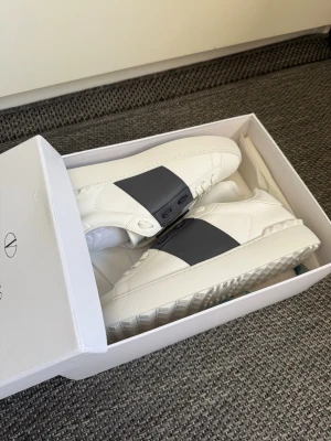 Valentino vita sneakers med svart band - Säljer ett par snygga vita sneakers från Valentino Garavani med ett brett svart band över vristen. Skorna har vita snören, diskret logga på plösen och coola nitar på sulan. Tillverkade i läder för en lyxig känsla och stilren look.