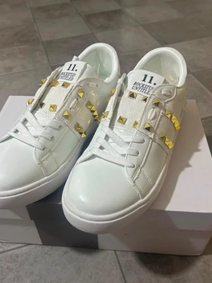 Vita sneakers med guldnitar Valentino - Snygga vita sneakers från Valentino med coola guldfärgade nitar längs sidorna och breda vita band. Skorna har en klassisk låg siluett, vita snören och detaljerad sula. Perfekta för dig som vill sticka ut med en exklusiv touch.