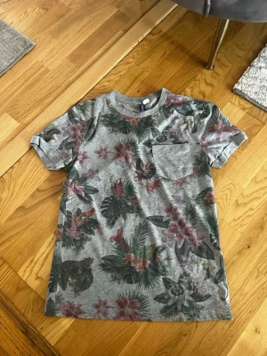 Grå t-shirt med blommigt tryck Eur XS - Snygg grå t-shirt med blommigt mönster i grönt, rött och rosa. T-shirten har en bröstficka och klassisk rund hals. Perfekt för dig som gillar en avslappnad sommar stil🔥☀️