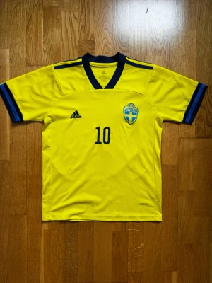 Sverige matchtröja Forsberg #10 Adidas - Gul matchtröja från Adidas för svenska landslaget med blå detaljer, nummer 10 och Forsberg på ryggen. Broderad landslagssköld på bröstet och klassisk V-ringning. Tillverkad i lätt och ventilerande funktionsmaterial, perfekt för fotboll.