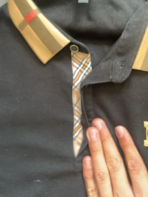 Svart Burberry skjorta med rutig krage - Säljer en svart skjorta från Burberry med klassisk beige och röd rutig krage och detaljer vid knäppningen. Skjortan har korta ärmar och en broderad logga på bröstet. Tillverkad i bomull och har en normal passform.