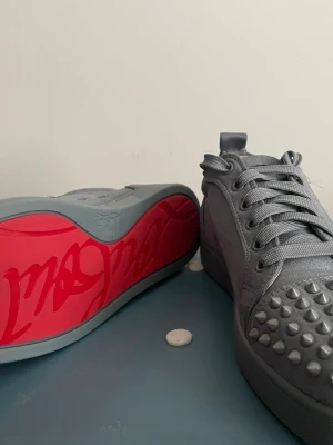 Gråa Louboutin sneakers med nitar - Säljer ett par gråa sneakers från Christian Louboutin med ikoniska röda sulor och coola nitar på tån. Skorna har en snygg matt finish och matchande grå skosnören. Perfekta för dig som vill sticka ut med en lyxig stil.