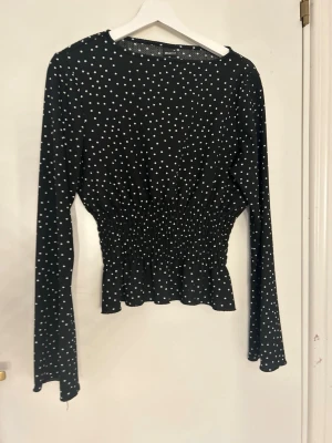 Svart prickig blus från Gina Tricot - Trendig svart blus från Gina Tricot med vita prickar och smockad midja som ger en snygg siluett. Blusen har långa ärmar och lätt utsvängd nederkant. Tillverkad i ett mjukt och stretchigt material som känns bekvämt hela dagen.
