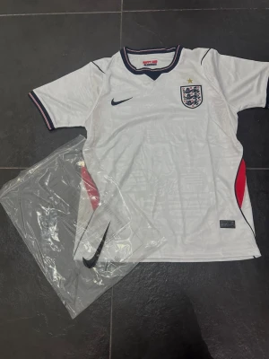 England Nike vit matchtröja - Säljer en vit England matchtröja från Nike med blå detaljer och röd accent vid sidorna. Tröjan har korta ärmar, broderad Nike-logga och Englands landslagsmärke på bröstet. Tillverkad i lätt och ventilerande material, perfekt för fotboll eller träning.