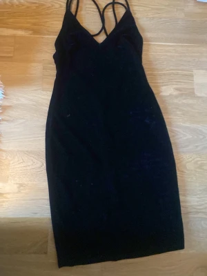 Svart sammetsklänning med smala band - Snygg svart klänning i sammet med smala axelband och v-ringad urringning. Klänningen har en kroppsnära passform och är ärmlös, perfekt för dig som gillar en stilren och elegant look.