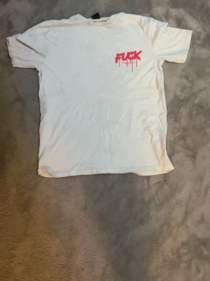Vit t-shirt med röd graffiti-text - Vit t-shirt med korta ärmar och stor röd graffiti-inspirerad text både fram och bak. Texten har en sprayad look med droppande effekt. Perfekt för dig som gillar streetwear och statement-plagg.