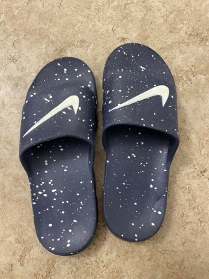 Blå Nike sandaler med vita stänk - Säljer ett par blå Nike sandaler med vita färgstänk och stor vit Swoosh-logga på ovandelen. Tillverkade i syntetmaterial med en bekväm, formad sula och unik undersida för extra grepp. Perfekta för chill dagar eller efter träningen.