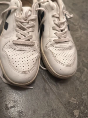Vita Veja sneakers med blå detalj - Snygga vita sneakers från Veja med blå V-logga på sidan och röd detalj vid hälen. Skorna har perforerad tå, vita snören och är tillverkade i läder. Klassisk låg modell som passar till det mesta.