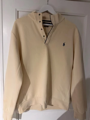 Beige långärmad tröja från Polo Ralph Lauren - Klassisk beige långärmad tröja från Polo Ralph Lauren med blå logga broderad på bröstet. Tröjan har knappar vid halsen och ribbade muddar vid ärmslut och nederkant. Perfekt för en clean och stilren look.