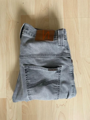 Grå jeans från Nudie Jeans - Säljer ett par grå jeans från Nudie Jeans med klassisk femficksmodell och raka ben. Jeansen har en snygg tvättad look, bruna detaljer vid midjan och är tillverkade i mjukt denimtyg. Perfekta för dig som gillar stilrena och tidlösa plagg.
