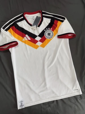 Tysklands fotbollströja Adidas - Säljer en vit Tyskland fotbollströja från Adidas med klassiskt mönster i svart, rött och gult över bröstet. Tröjan har rund hals med röd kant, svarta och vita ränder på axlarna samt Tysklands landslagsemblem på bröstet. Tillverkad i lätt och ventilerande material.